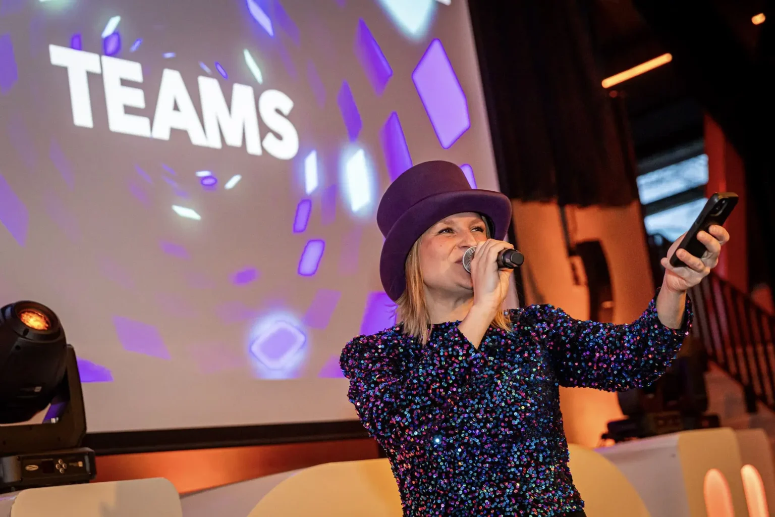 Vrouwelijke quizmaster met paarse hoed presenteert interactieve pubquiz voor teams, met kleurrijke projecties op de achtergrond.