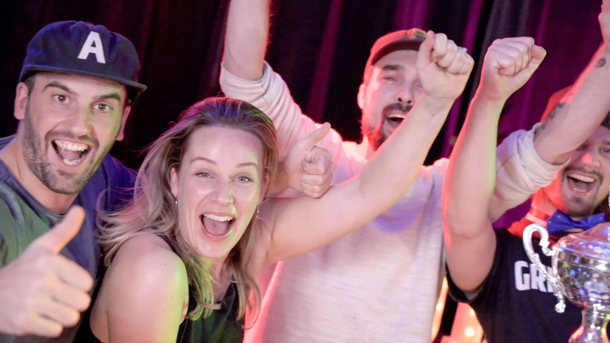 Groep mensen viert uitbundig een overwinning met een beker en kleurrijke lichten op de achtergrond. Perfect moment voor een teambuilding evenement met een ingehuurde quizmaster.