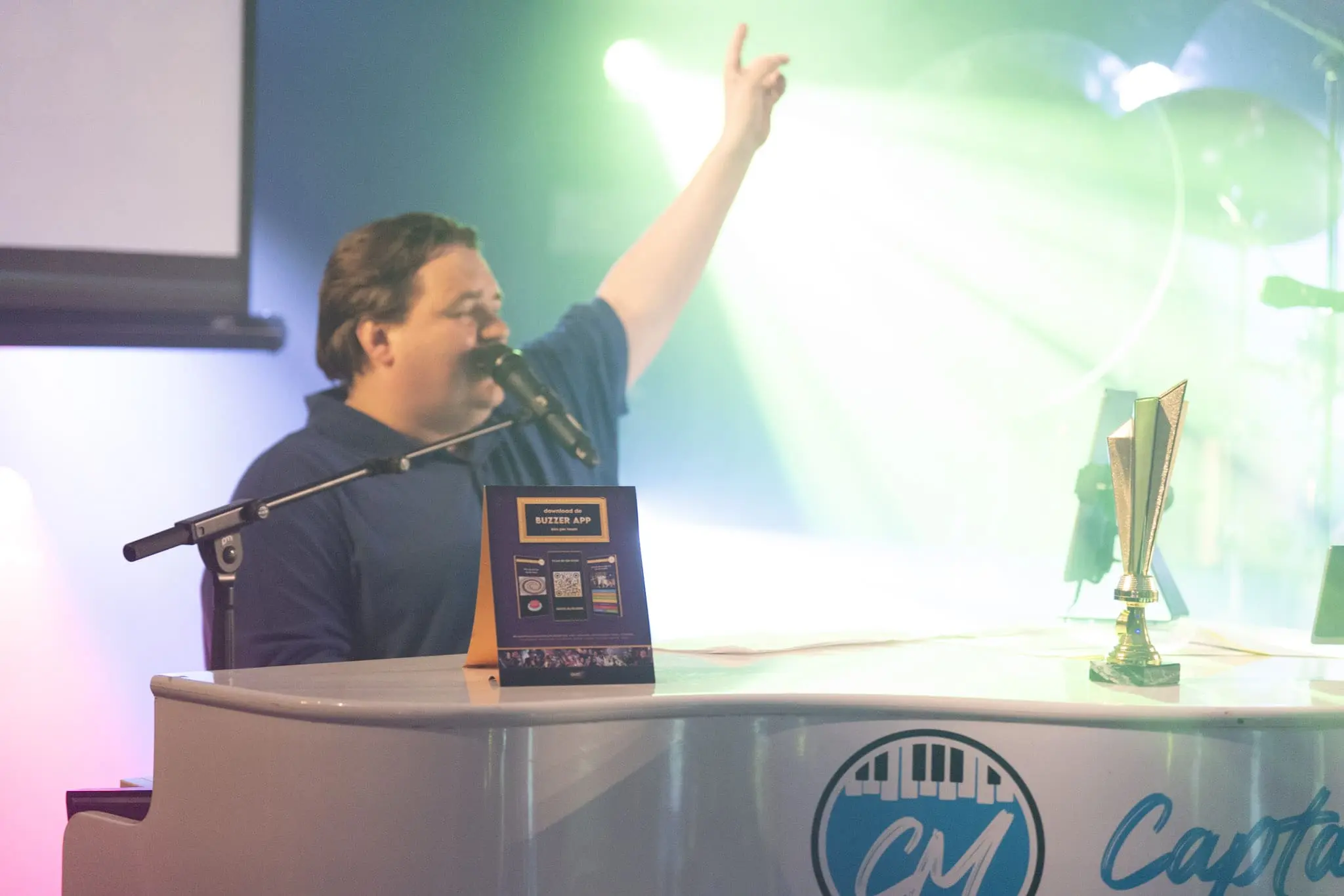 Quizmaster presenteert levendige quizshow met microfoon, trofee en buzzer-app promotie in dynamische sfeer.