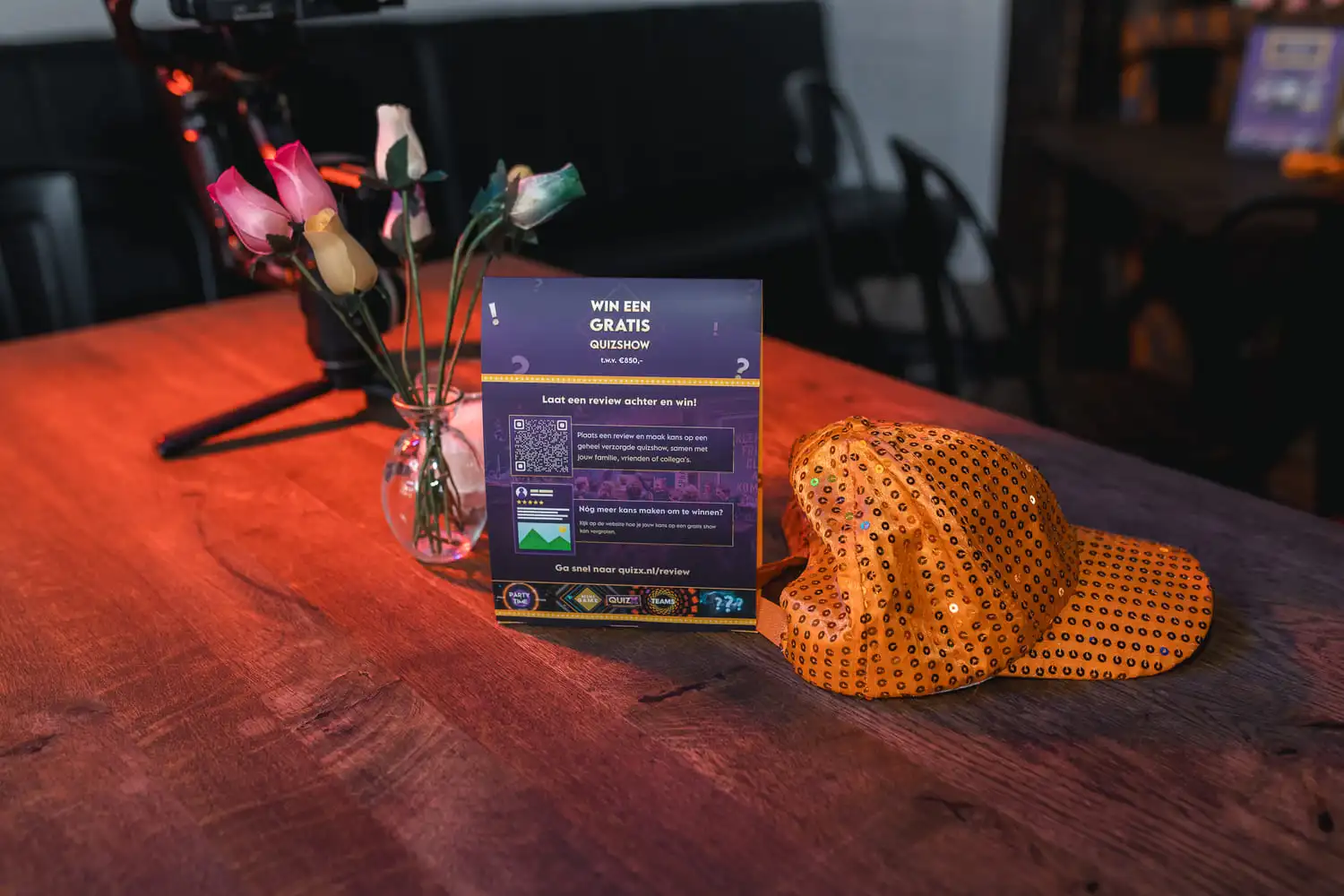 Feestelijke tafelopstelling met oranje glitterpet en promotiebord voor gratis quizshow deelname, kleurrijke kunstrozen in vaas.